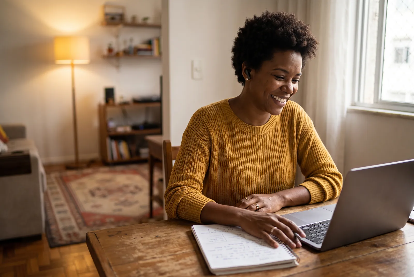 Corretora em casa, com fones de ouvido, sorrindo enquanto acompanha um encontro ao vivo no notebook